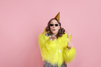 Photo of Birthday celebration. Woman with glass of sparkling wine blowing confetti on pink background