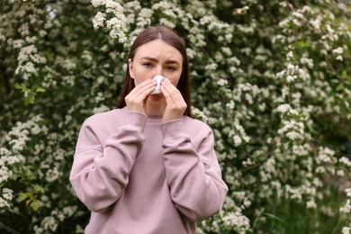 Photo of Woman with paper tissue suffering from seasonal pollen allergy near blossoming shrub on spring day