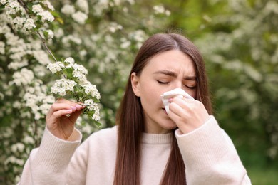 Photo of Woman with paper tissue suffering from seasonal pollen allergy near blossoming shrub on spring day