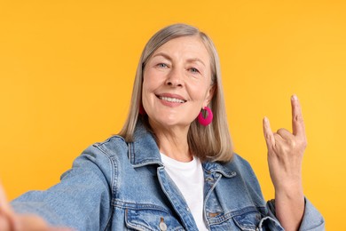 Photo of Senior woman in stylish outfit making rock gesture while taking selfie on orange background