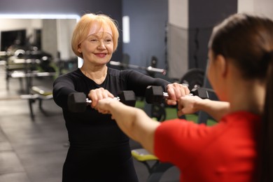 Photo of Senior woman exercising with dumbbells supervised by professional trainer in gym, selective focus