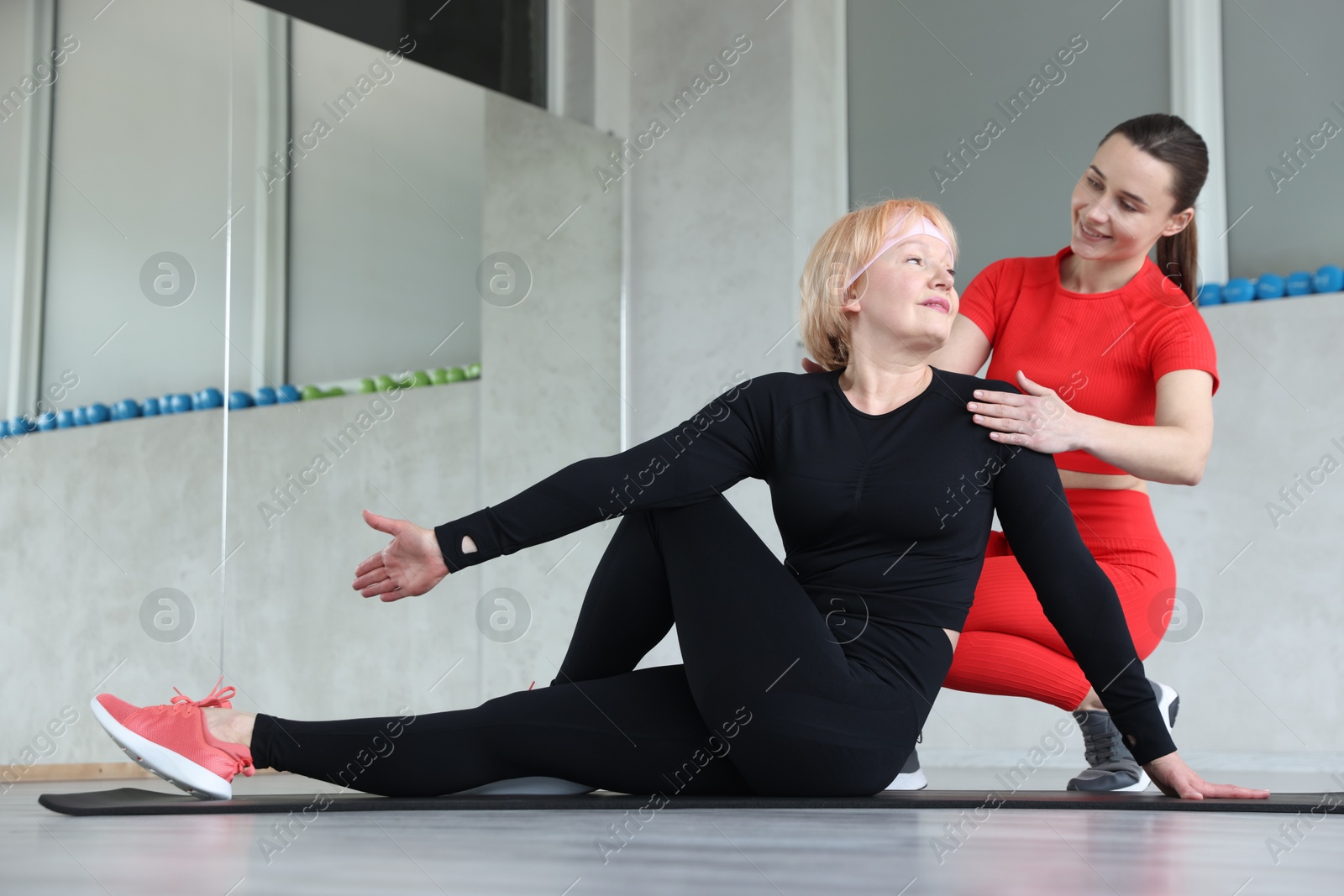Senior woman exercising on yoga mat supervised by professional trainer in gym Photo of Senior woman exercising on yoga mat supervised by professional trainer in gym