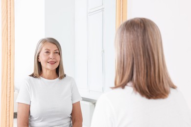 Photo of Beautiful senior woman looking at herself in mirror indoors