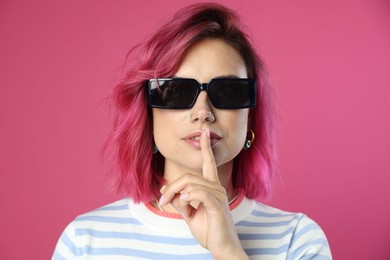 Photo of Beautiful woman with bright dyed hair in sunglasses making hush gesture on pink background