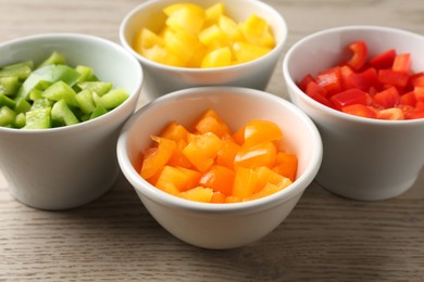 Photo of Pieces of fresh colorful bell peppers in bowls on wooden table, closeup