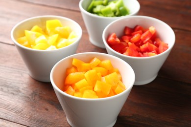 Photo of Pieces of fresh colorful bell peppers in bowls on wooden table, closeup