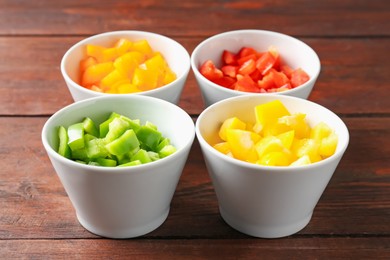 Photo of Pieces of fresh colorful bell peppers in bowls on wooden table, closeup