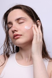 Photo of Beautiful woman applying skincare product onto her face on light grey background, closeup