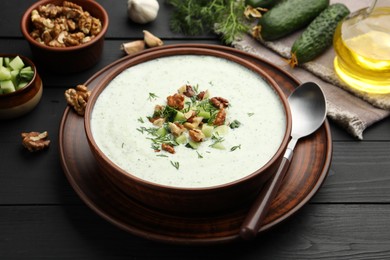Photo of Tasty cucumber soup with dill and walnuts served on black wooden table, closeup