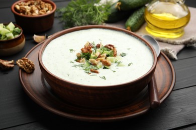 Photo of Tasty cucumber soup with dill and walnuts served on black wooden table, closeup