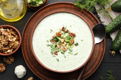 Photo of Tasty cucumber soup with dill and walnuts served on black wooden table, flat lay