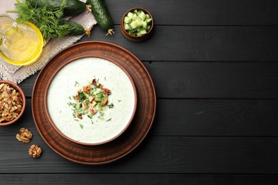 Photo of Tasty cucumber soup with dill, walnuts and ingredients on black wooden table, flat lay. Space for text