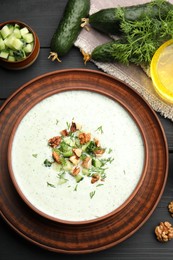 Photo of Tasty cucumber soup with dill, walnuts and ingredients on black wooden table, flat lay