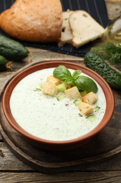 Photo of Tasty cucumber soup with croutons, basil, bread and vegetables on wooden table, closeup