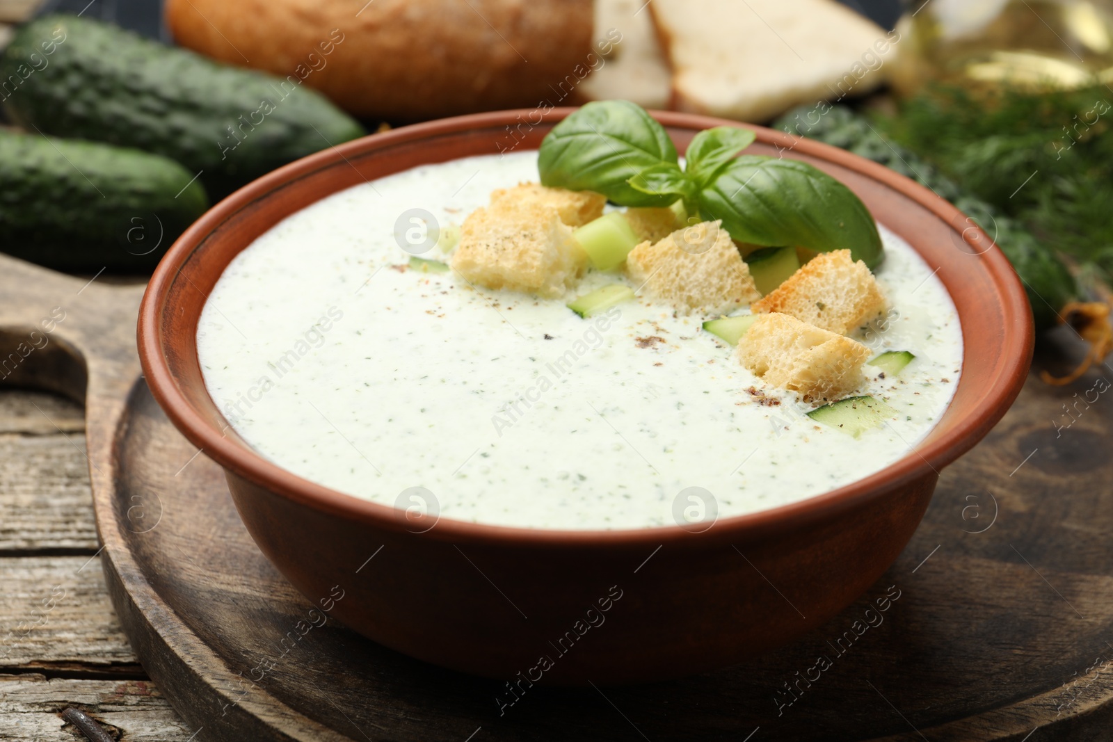 Photo of Tasty cucumber soup with croutons, basil, bread and vegetables on wooden table, closeup