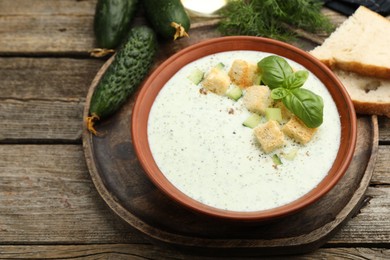 Photo of Tasty cucumber soup with croutons, basil, bread and vegetables on wooden table, closeup
