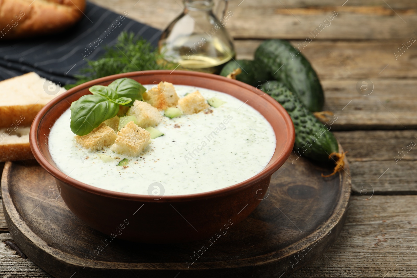 Photo of Tasty cucumber soup with croutons, basil, bread and vegetables on wooden table, closeup