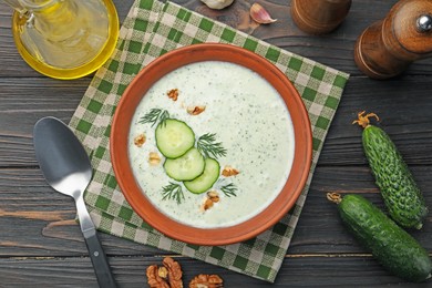 Photo of Tasty cucumber soup with dill, walnuts, spoon and ingredients on wooden table, flat lay