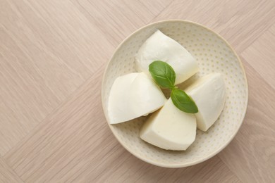 Photo of Pieces of delicious mozzarella cheese and basil in bowl on wooden table, top view. Space for text
