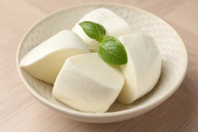 Photo of Pieces of delicious mozzarella cheese and basil in bowl on wooden table, closeup