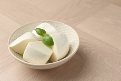 Photo of Pieces of delicious mozzarella cheese and basil in bowl on wooden table, closeup