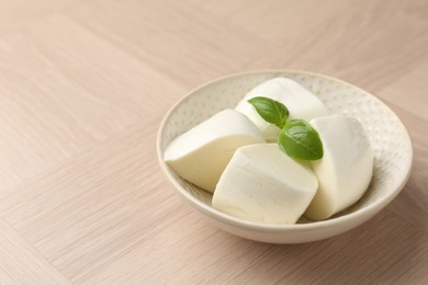 Photo of Pieces of delicious mozzarella cheese and basil in bowl on wooden table, closeup. Space for text