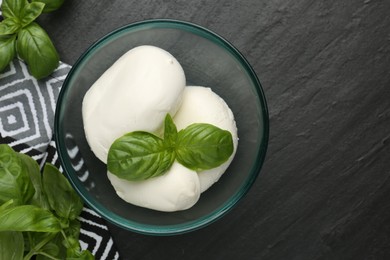 Photo of Pieces of delicious mozzarella cheese and basil in bowl on dark textured table, flat lay. Space for text