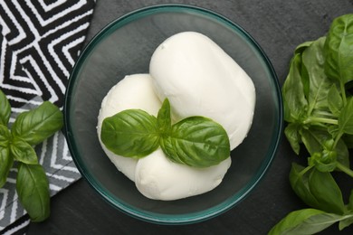 Photo of Pieces of delicious mozzarella cheese and basil in bowl on dark textured table, flat lay