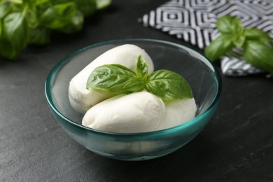 Photo of Pieces of delicious mozzarella cheese and basil in bowl on dark textured table, closeup