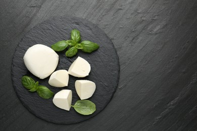 Photo of Pieces of delicious mozzarella cheese and basil on dark textured table, top view. Space for text