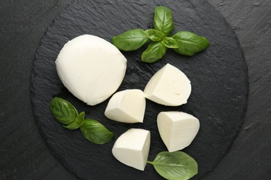 Photo of Pieces of delicious mozzarella cheese and basil on dark textured table, top view