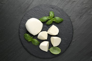 Photo of Pieces of delicious mozzarella cheese and basil on dark textured table, top view
