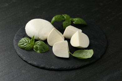 Photo of Pieces of delicious mozzarella cheese and basil on dark textured table, closeup