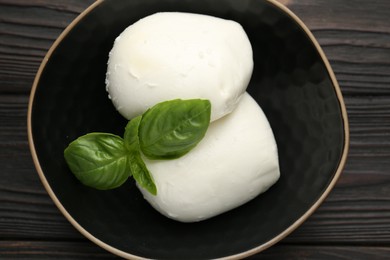Photo of Pieces of delicious mozzarella cheese and basil in bowl on wooden table, top view