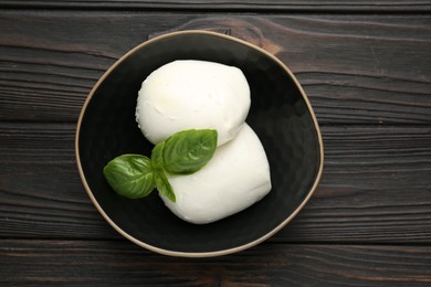 Photo of Pieces of delicious mozzarella cheese and basil in bowl on wooden table, top view