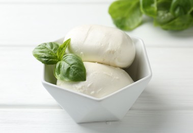 Photo of Pieces of delicious mozzarella cheese and basil in bowl on white wooden table, closeup