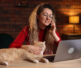 Photo of Smiling woman talking by smartphone and stroking cat at desk in home office
