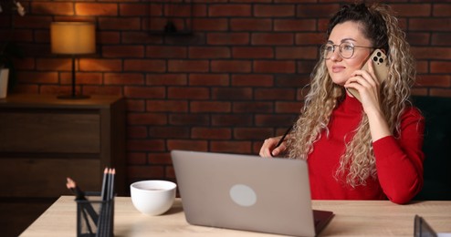 Photo of Woman talking by smartphone at desk with laptop in home office