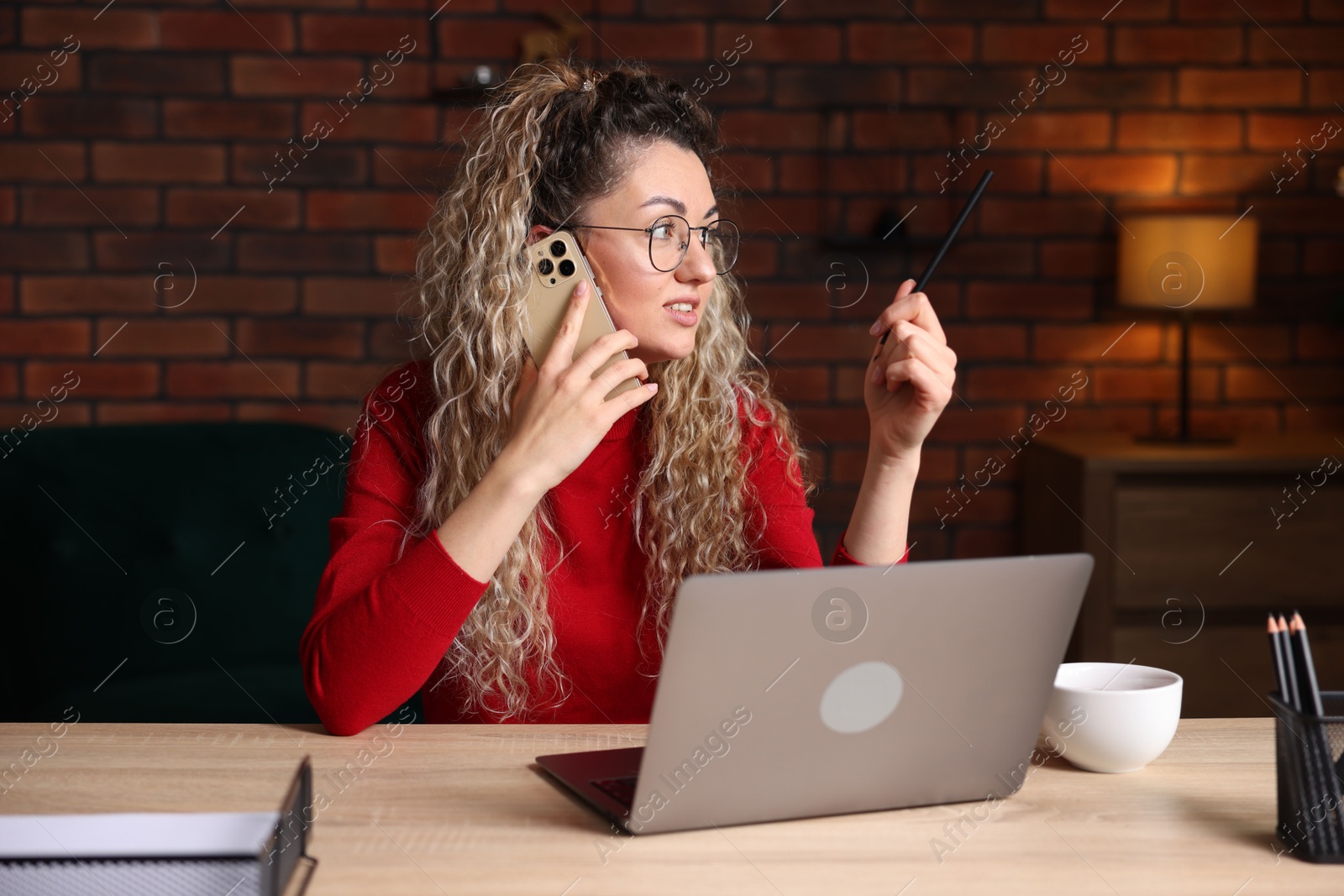 Woman talking by smartphone at desk with laptop in home office Photo of Woman talking by smartphone at desk with laptop in home office