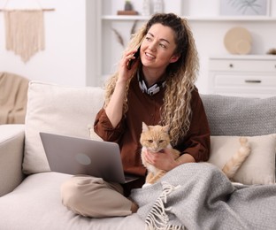 Photo of Smiling woman talking by smartphone with laptop and cat on sofa in home office