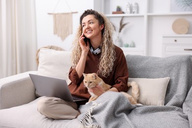 Photo of Smiling woman talking by smartphone with laptop and cat on sofa in home office
