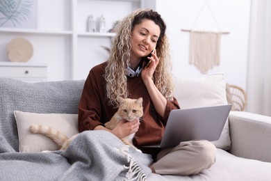 Photo of Smiling woman talking by smartphone with laptop and cat on sofa in home office