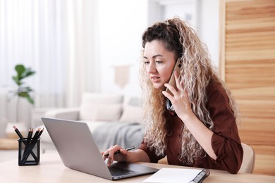 Photo of Woman talking by smartphone at desk with laptop in home office