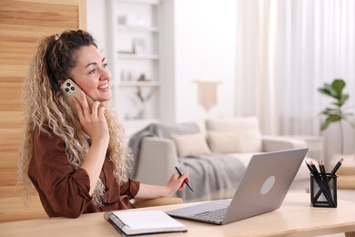 Photo of Smiling woman talking by smartphone at desk with laptop in home office