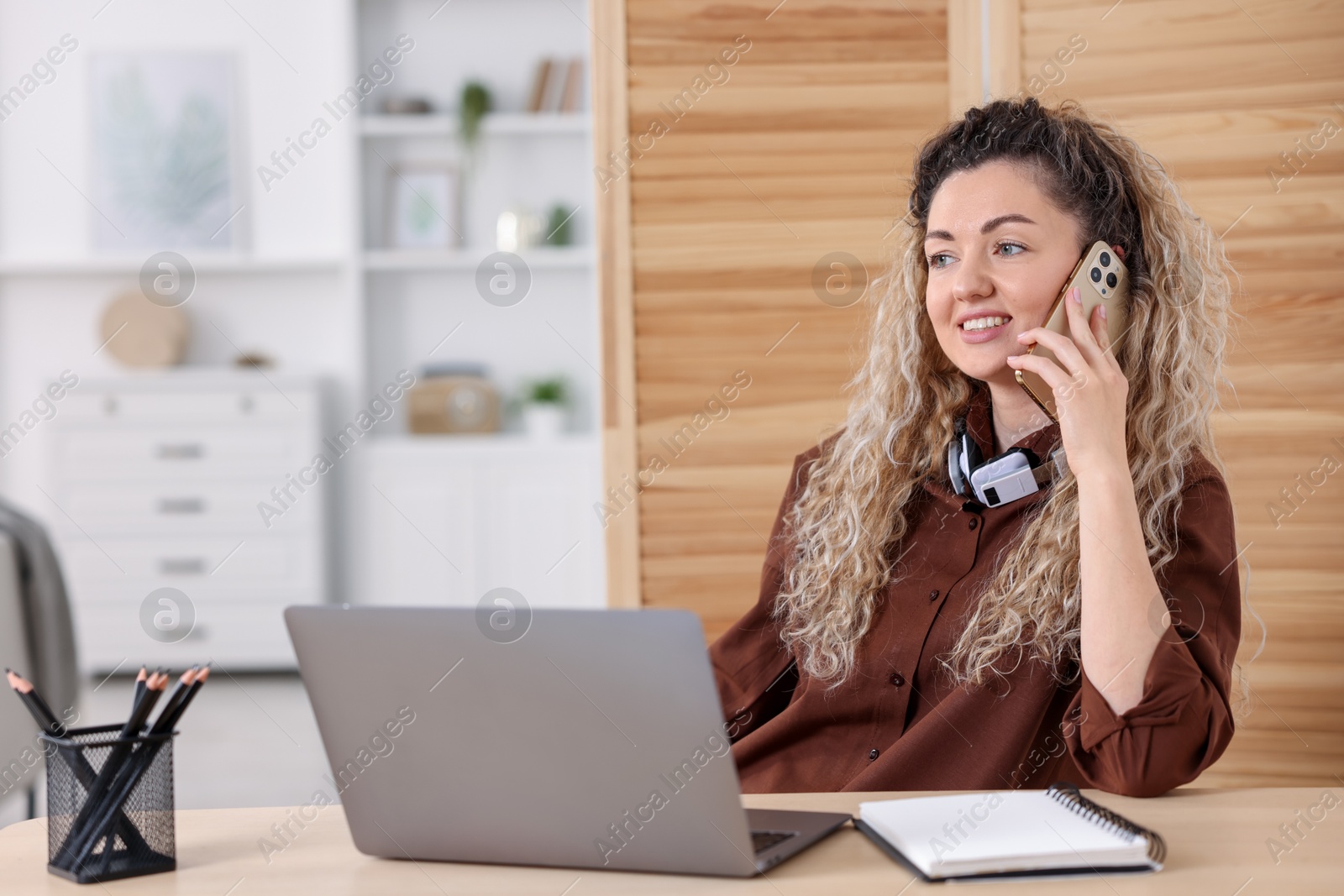 Smiling woman talking by smartphone at desk with laptop in home office Photo of Smiling woman talking by smartphone at desk with laptop in home office