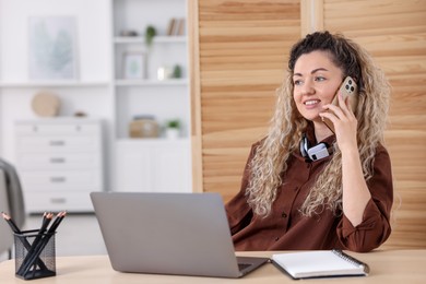 Photo of Smiling woman talking by smartphone at desk with laptop in home office