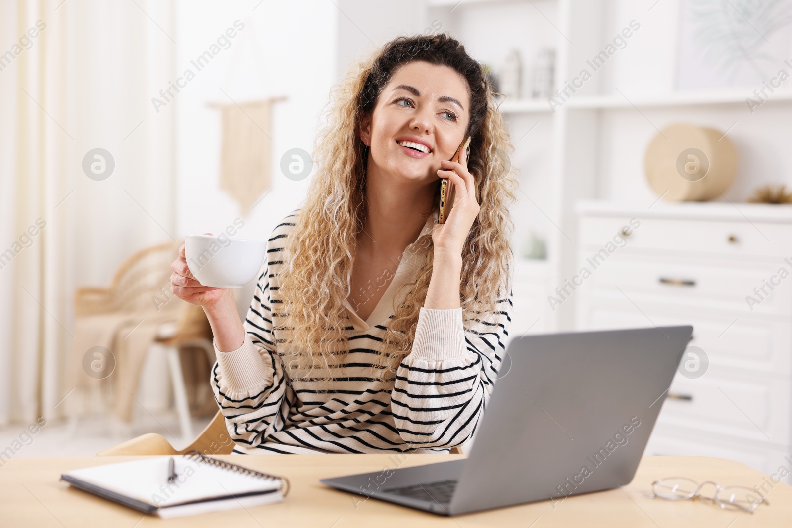 Smiling woman talking by smartphone at desk with laptop in home office Photo of Smiling woman talking by smartphone at desk with laptop in home office