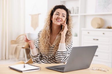 Photo of Smiling woman talking by smartphone at desk with laptop in home office