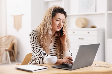Photo of Woman talking by smartphone and working with laptop at desk in home office