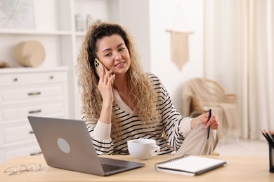 Photo of Smiling woman talking by smartphone at desk with laptop in home office